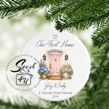 A personalized round white Christmas ornament hanging from green pine branches. The ornament features a printed illustration of a pink front door with flowers, and custom text reading 'Our First Home', 'Gray & Sally', and '6 Clenside Street Palmside 2026'.