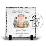 Personalized square slate memorial plaque on a black display stand, featuring a watercolor pink front door, potted flowers, and custom text reading 'Our First Home' and 'Greg & Sally' with address and year.